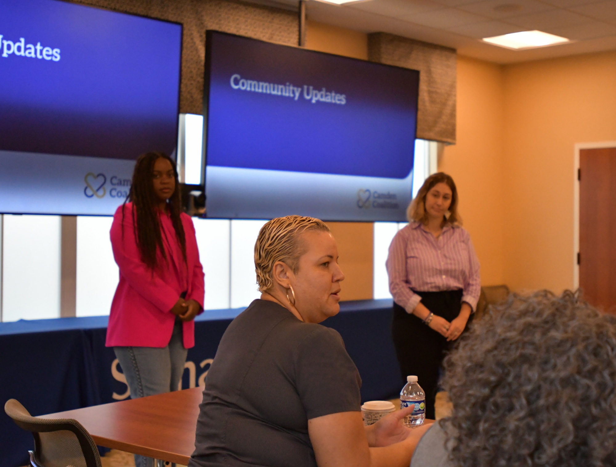 A person with short blonde hair faces a room as she's speaking. In the background, two Camden Coalition staff members stand in front of a large screen reading "Community updates" as they listen